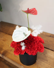 Load image into Gallery viewer, A floral arrangement featuring bright red carnations, a white anthurium, and a red anthurium stem, styled in a black pot on a wooden surface against a neutral wall.