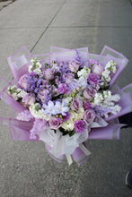 Load image into Gallery viewer, A bouquet of purple, lavender, and white flowers wrapped in translucent light purple paper and a white ribbon. 
